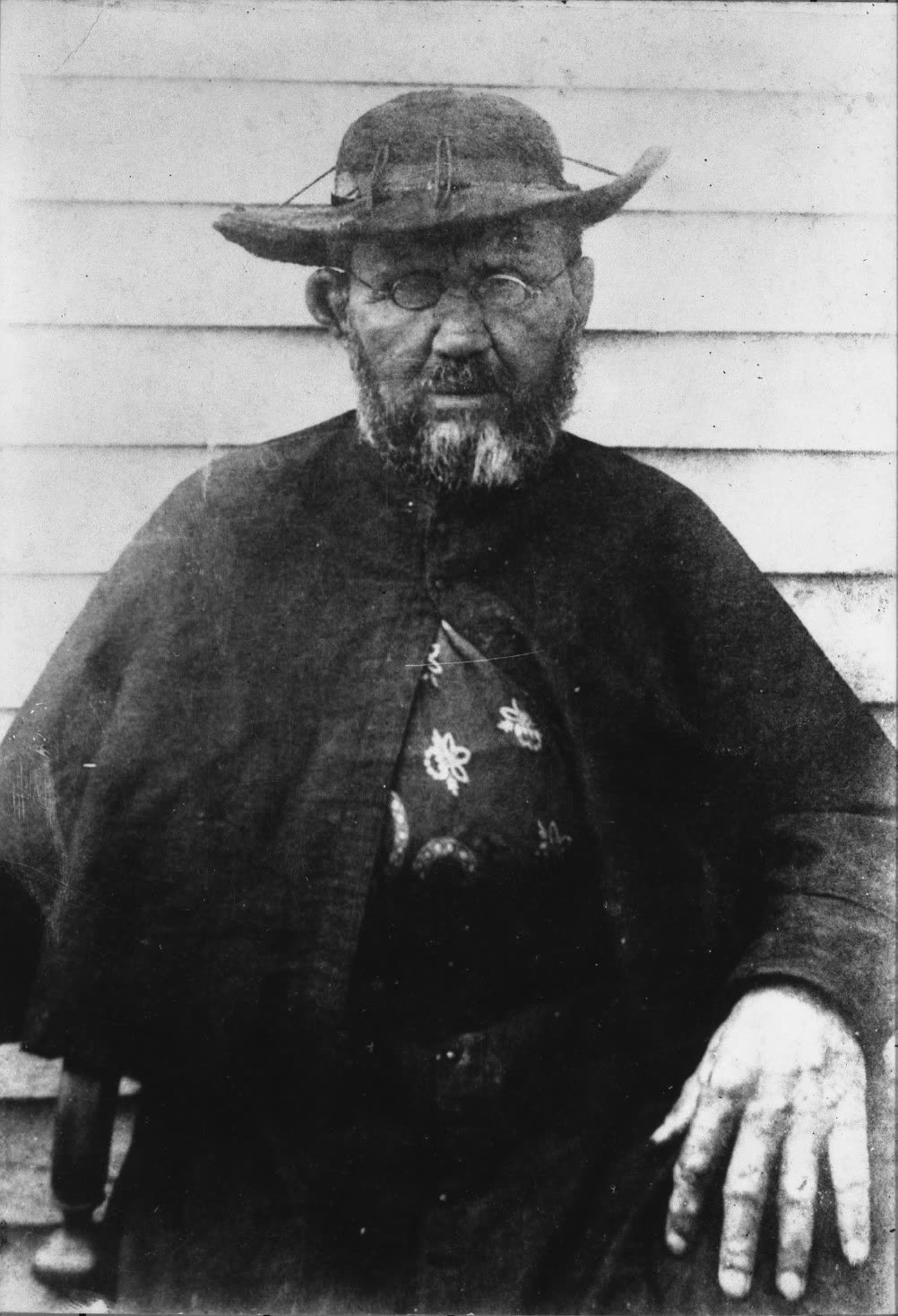 Black-and-white photograph of Fr. Damien de Veuster in his final year, a bearded priest in simple dark clothing.
